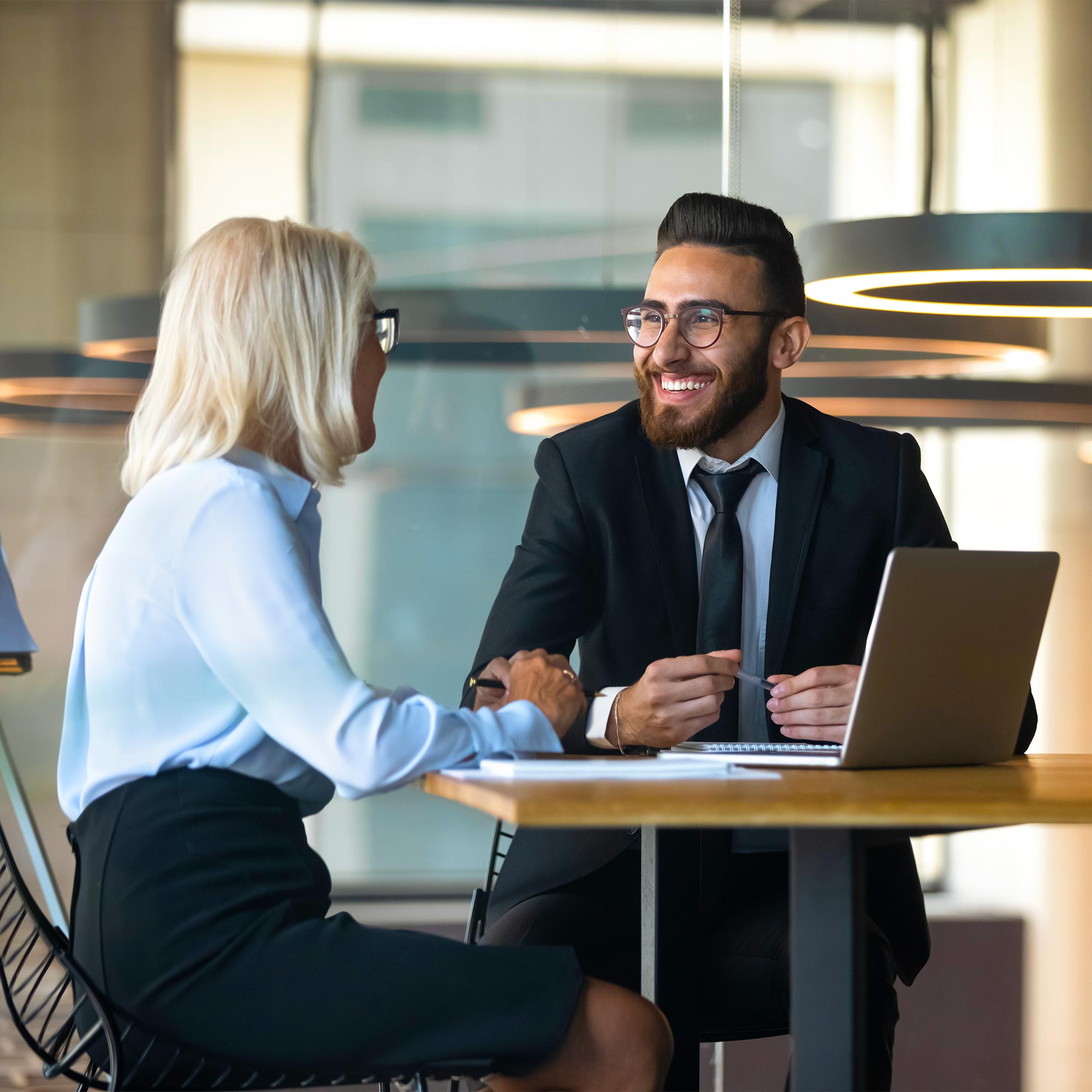 A man and woman sit at a table with a laptop and are having a discussion.
